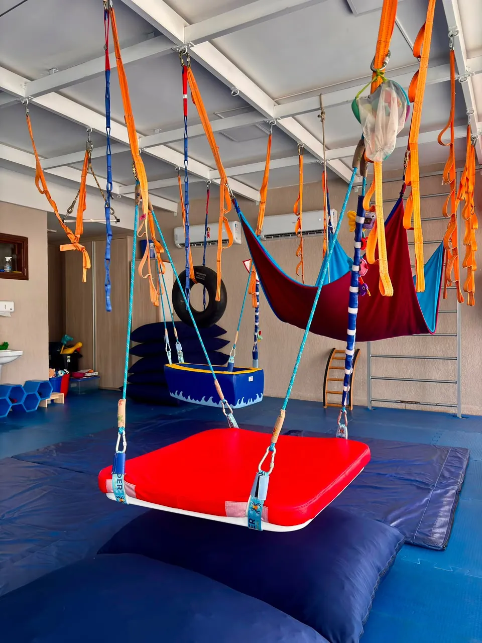 Balanços da sala de Integração Sensorial: plataforma, balanço-barco, rede e pneu suspensos sobre o tatame azul.